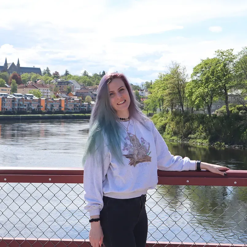 Foto von mir, wo ich auf einer Brücke stehe und in die Kamera lächle. Hinter mir ist ein Fluss und eine Häuserfront in Trondheim, Norwegen, zu sehen.